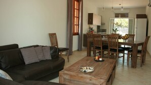 A living room with a dark brown sofa, wooden dining table, and chairs. A kitchen is visible in the background with wooden cabinets and a refrigerator. - House / Villa - Port-Vecchio (Porto-Vecchio)