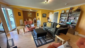 A living room with a sofa, armchair, and ottoman, a bookshelf, and a dining area with a table and chairs. - Excellent ground floor 2 Bed apt overlooking golf course with private parking. (Watton)