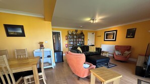 A living room with a wooden dining table, a sofa, a chair, and a small table. There is a bookshelf and a door in the background. - Excellent ground floor 2 Bed apt overlooking golf course with private parking. (Watton)