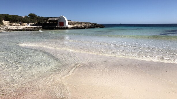 Une plage aux eaux claires et peu profondes, un bâtiment blanc avec une porte rouge, et un paysage rocheux en arrière-plan.