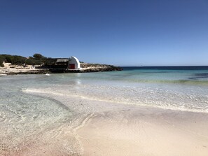 Une plage aux eaux claires et peu profondes, un bâtiment blanc avec une porte rouge, et un paysage rocheux en arrière-plan.