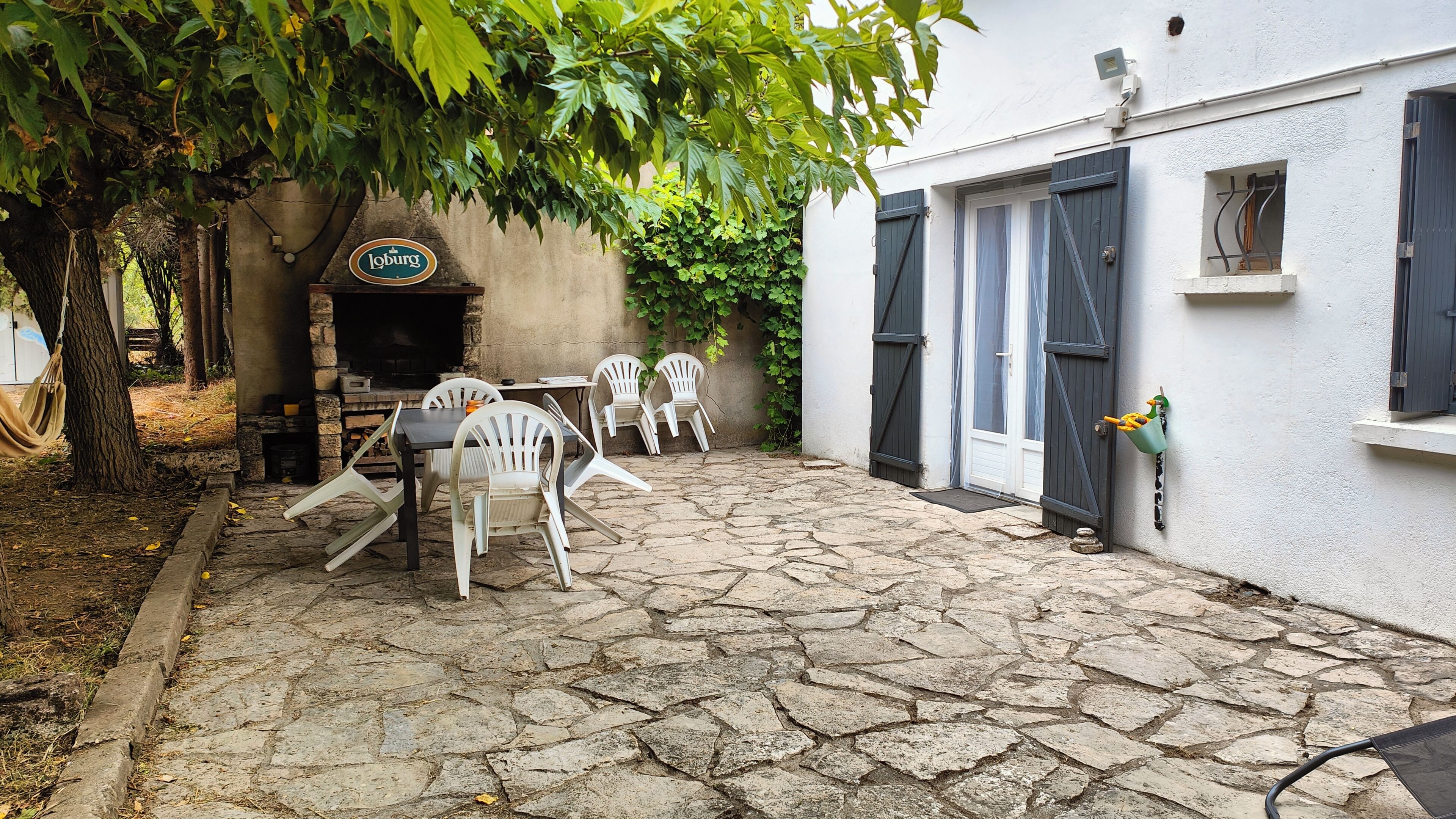 A stone-paved courtyard with outdoor seating, a stone fireplace, and a white building with shutters.