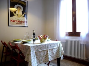 A dining table with a floral tablecloth, a bowl of fruit, wine glasses, and a bottle of wine. There is a poster on the wall and a window with curtains.