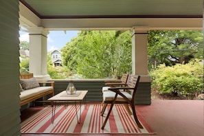 A covered porch with a seating area, a table, and a view of a house and greenery.