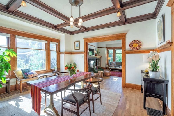 A spacious dining area with a long wooden table, multiple chairs, and a bench. There are large windows providing natural light and views of greenery outside.