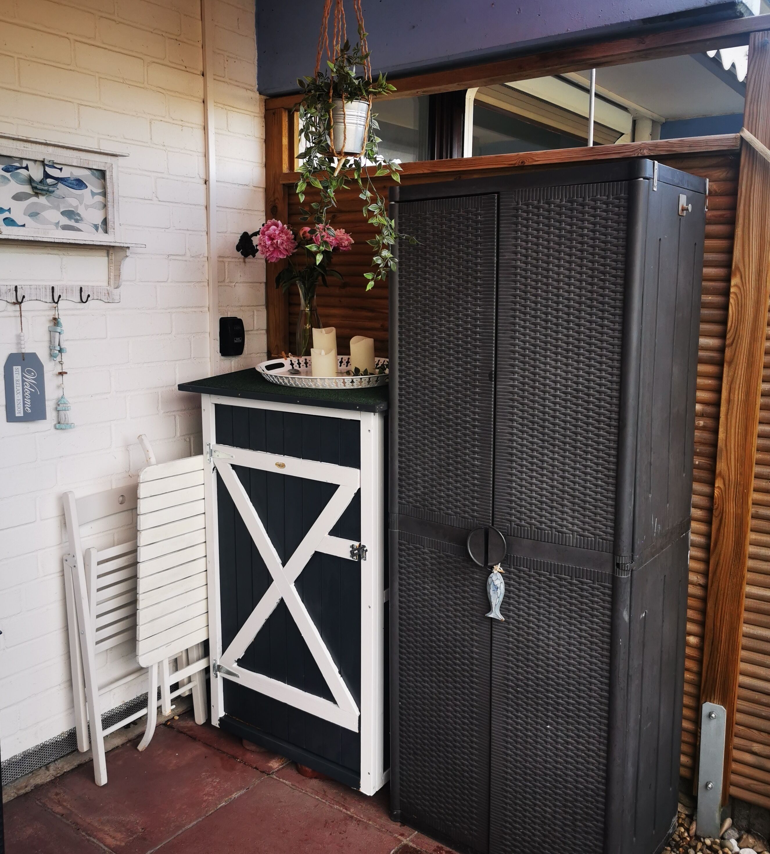 A compact outdoor area with a black metal cabinet, a white bench, and a small table with candles and flowers.