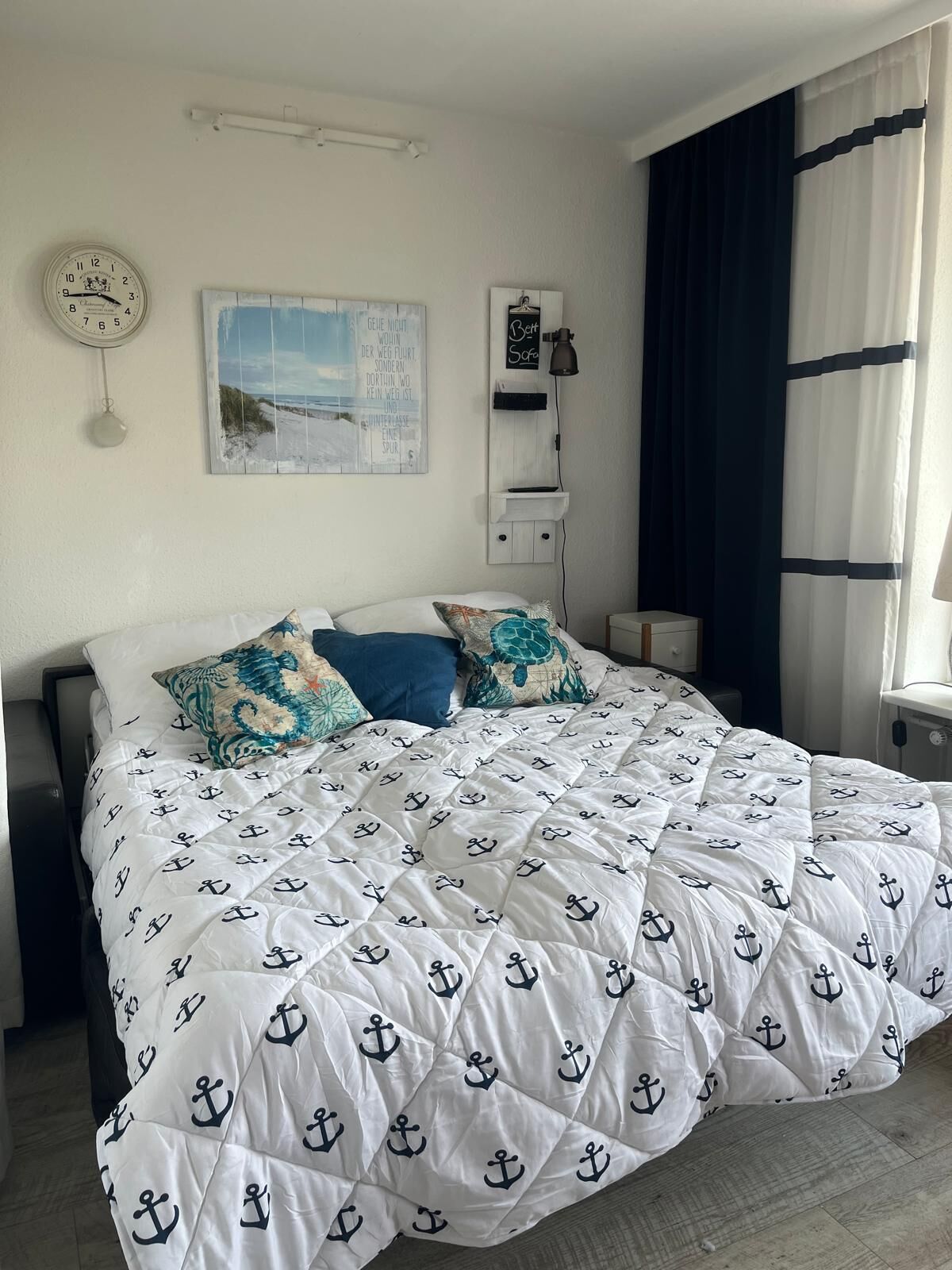 Een net opgemaakt bed met een witte deken met ankerprint, een wandklok, een ingelijste foto en een bedlampje.