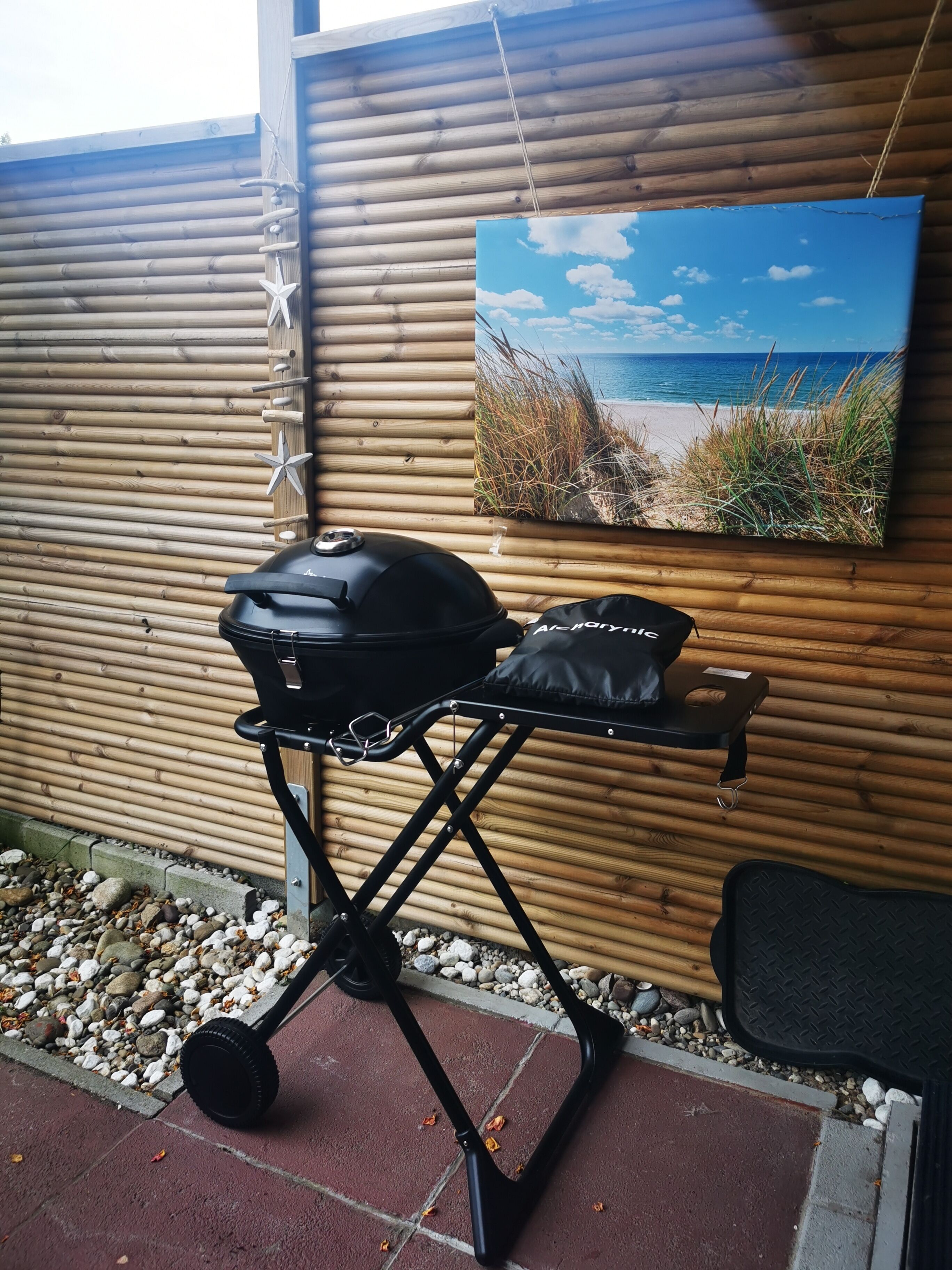 Ein tragbarer Grill mit schwarzem Deckel und schwarzem Tablett, auf einer Betonfläche mit Holzzaun im Hintergrund.