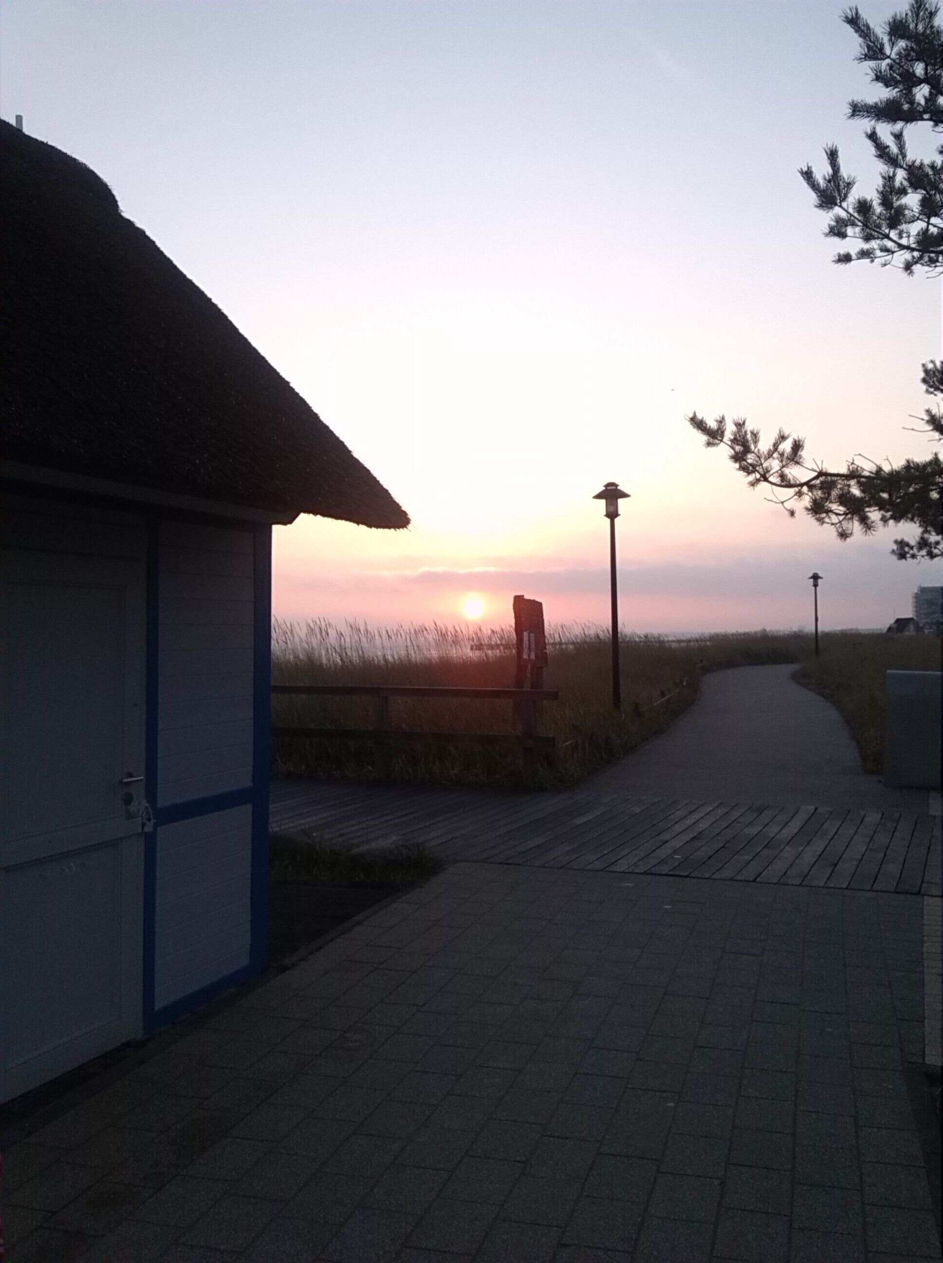 Ein Pfad führt zu einem Gebäude mit Strohdach, einer Straßenlaterne und Blick auf den Sonnenuntergang.