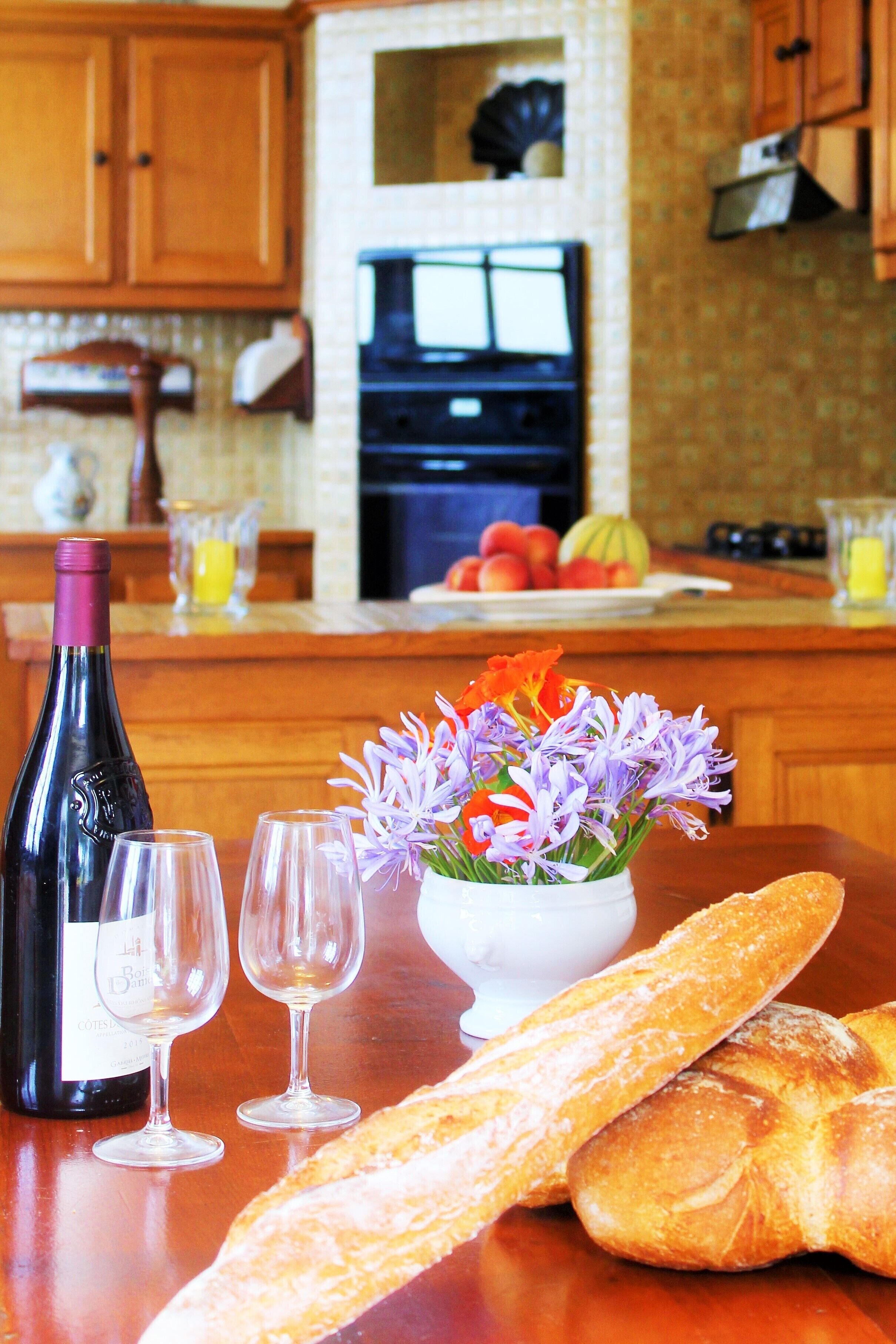 A kitchen counter with a bottle of wine, two glasses, a vase with flowers, and a loaf of bread.