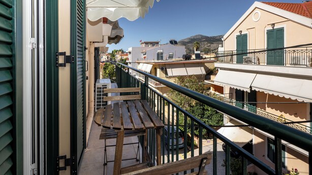 Ein Balkon mit einer Holzbank, einem Geländer und Blick auf benachbarte Gebäude und einen Berg.