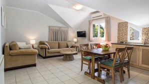 Ein gemütliches Wohnzimmer mit zwei Sofas, einem Couchtisch und einem Esstisch mit Stühlen. An der Wand ist ein Fernseher angebracht und es gibt ein Fenster mit Vorhängen.