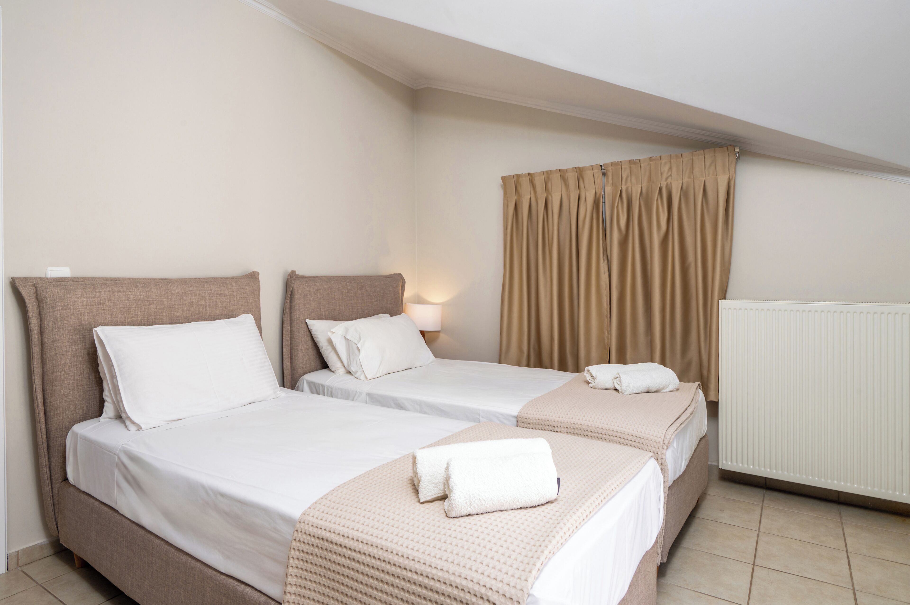A hotel room with two single beds, beige bedding, a radiator, and a window with beige curtains.