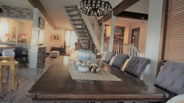Un espace repas avec une longue table en bois, des chaises rembourrées et un élément décoratif central. Un escalier avec une rampe est visible à l’arrière-plan.