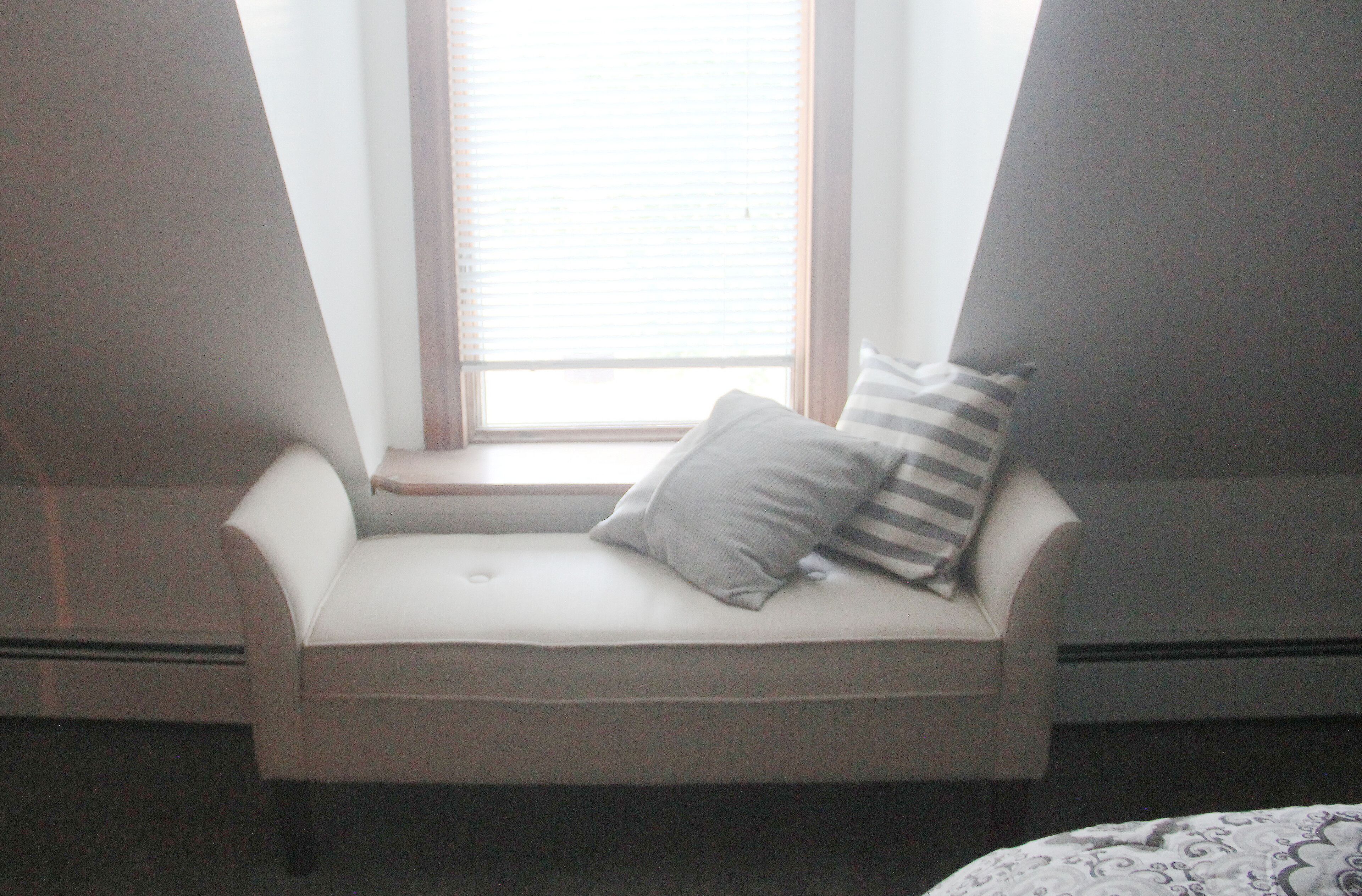 A corner seating area with a cushioned bench, a window with blinds, and two pillows.
