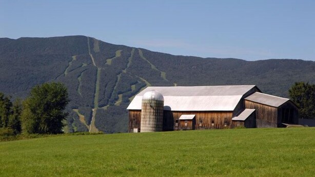 A rustic barn with a silo, surrounded by green fields and mountains in the background.