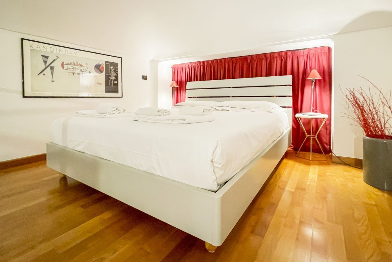 A bedroom with a white bed, red curtains, a small table with a lamp, and a framed picture on the wall.
