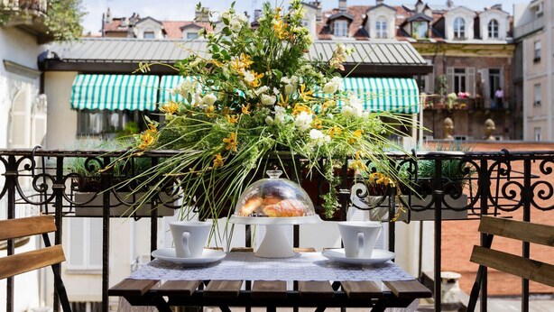 A table with a floral centerpiece, two white cups, and a glass dome with pastries on a balcony overlooking a cityscape. - LuxRent Loft Lagrange Sauna Gym (Torino)