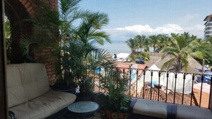 Een balkon met een bank en een kleine tafel, met uitzicht op een strand met palmbomen en een gebouw op de achtergrond.