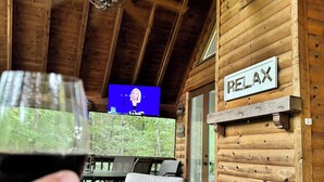 Ein Interieur einer Holzhütte mit Deckenventilator, einem 'RELAX'-Schild, einem Essbereich mit Tisch und Stühlen sowie einem Glas Rotwein.