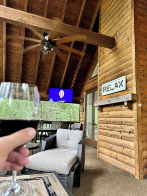 L’intérieur d’un chalet en bois, avec un ventilateur de plafond, une pancarte indiquant « RELAX », un coin repas avec une table et des chaises, et un verre de vin rouge.