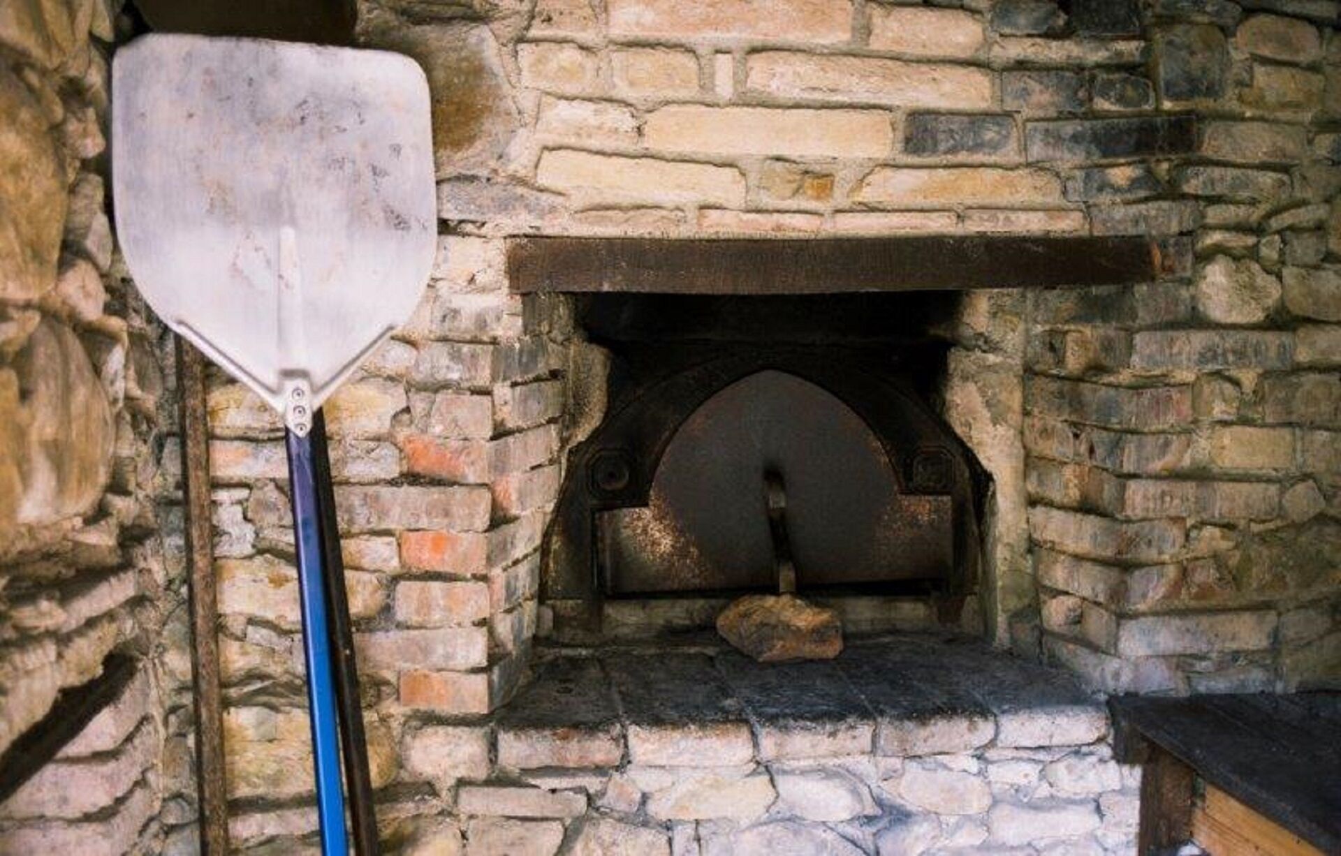 A stone fireplace with a metal door, a shovel, and a wooden bench.
