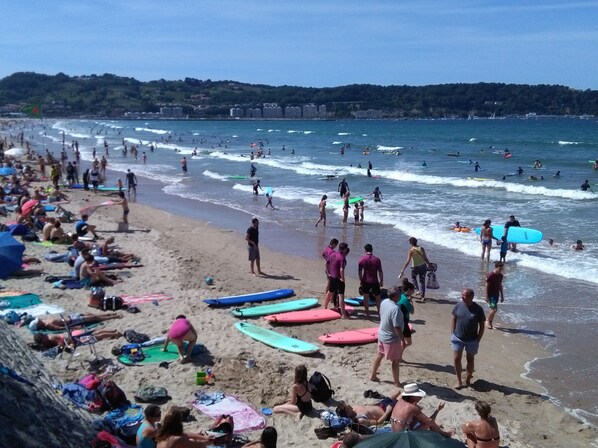 Une plage bondée où les gens se prélassent au soleil et se baignent, et où des planches de surf sont alignées sur le sable.