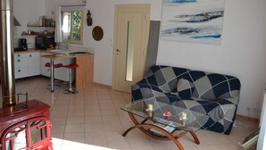 A living room with a glass coffee table, a sofa, and a kitchen area with a stove and bar stools. - Air-conditioned detached house 60m2 with garden (Sénas)