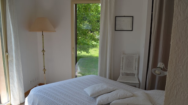 A bedroom with a large bed, a bedside lamp, a chair, and a view of a garden. - Air-conditioned detached house 60m2 with garden (Sénas)