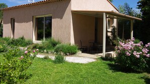 A small, single-story house with a covered patio, large windows, and a garden with pink flowers. - Air-conditioned detached house 60m2 with garden (Sénas)