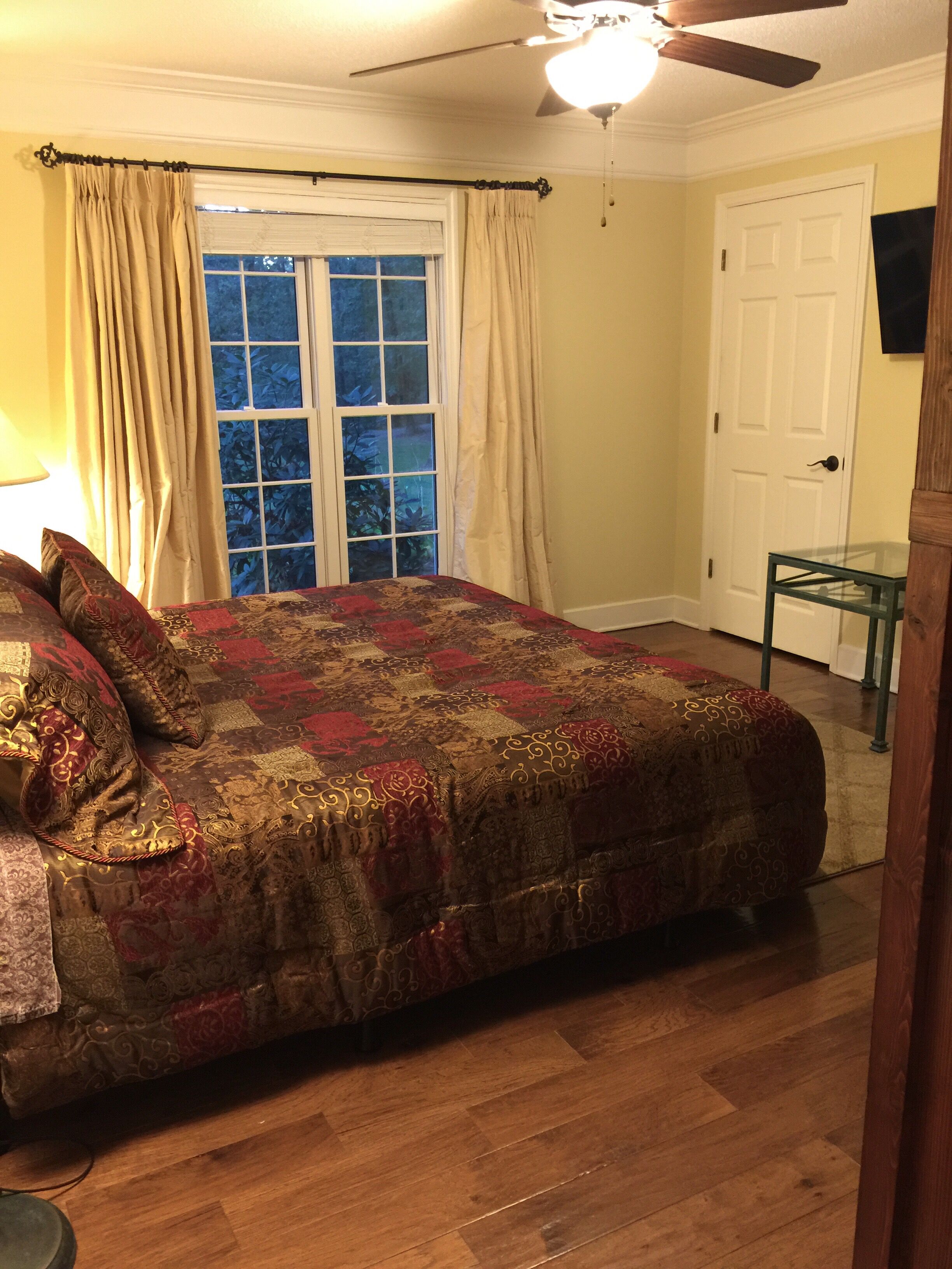 A bedroom with a large bed, a ceiling fan, a window with curtains, a door, and a small table.