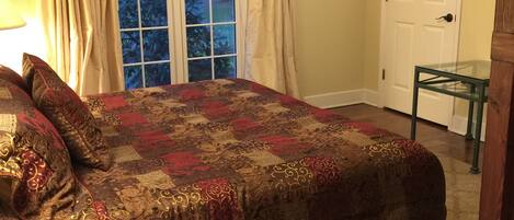 A bedroom with a large bed, a ceiling fan, a window with curtains, a door, and a small table.