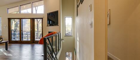 A spacious room with a wooden floor, a staircase with a railing, and a large window overlooking a deck.