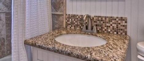A bathroom with a granite countertop sink, a mirror with lights, a shower curtain, and a tiled wall.
