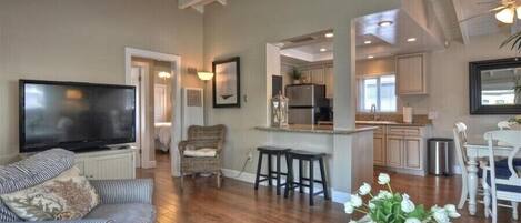 A spacious living area with a large flat-screen TV, a comfortable sofa, a dining table with chairs, and a kitchen area with a refrigerator and cabinets.