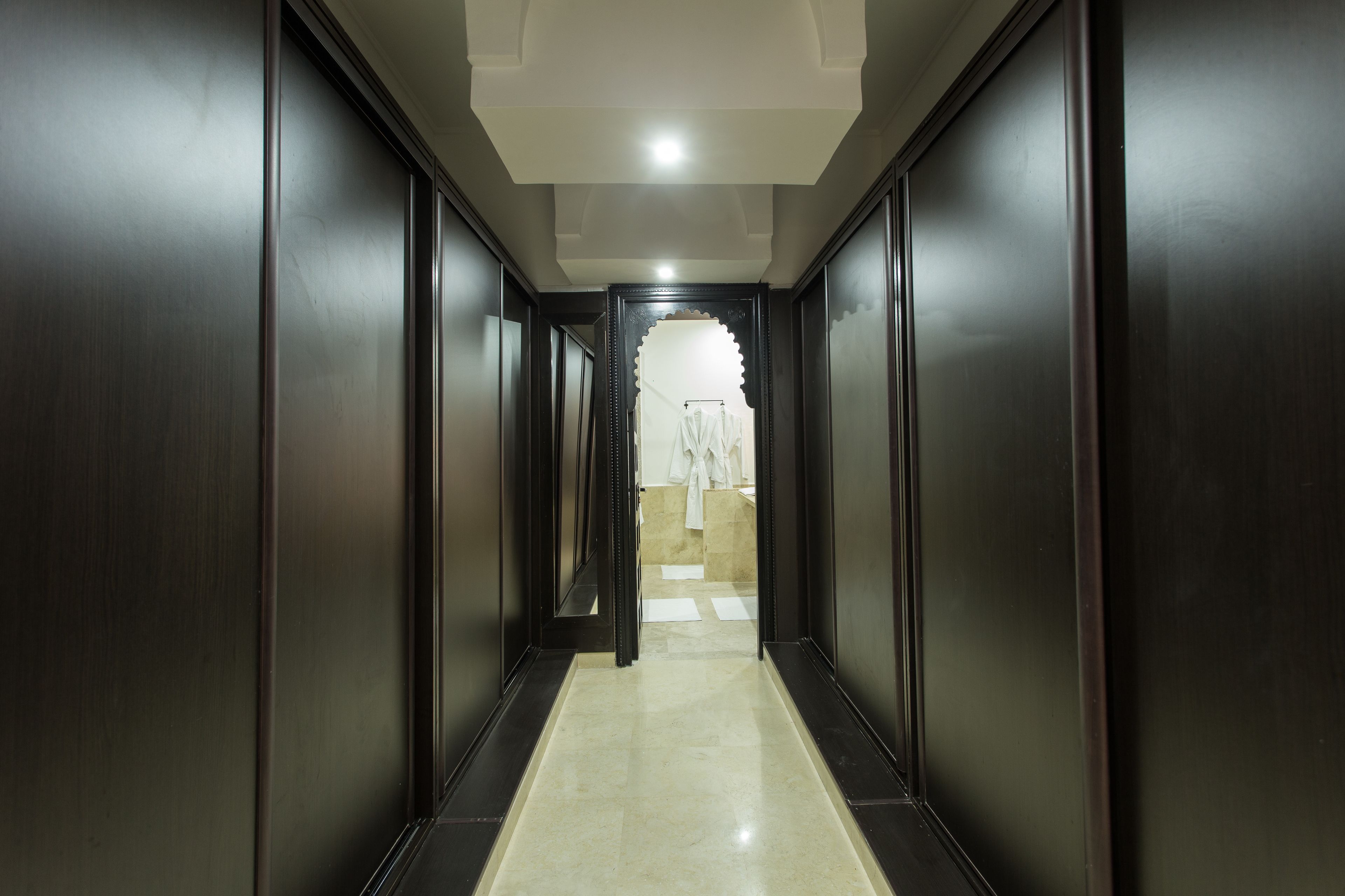 A narrow, dark wooden hallway with a tiled floor leading to a bathroom with a white sink and a towel rack.