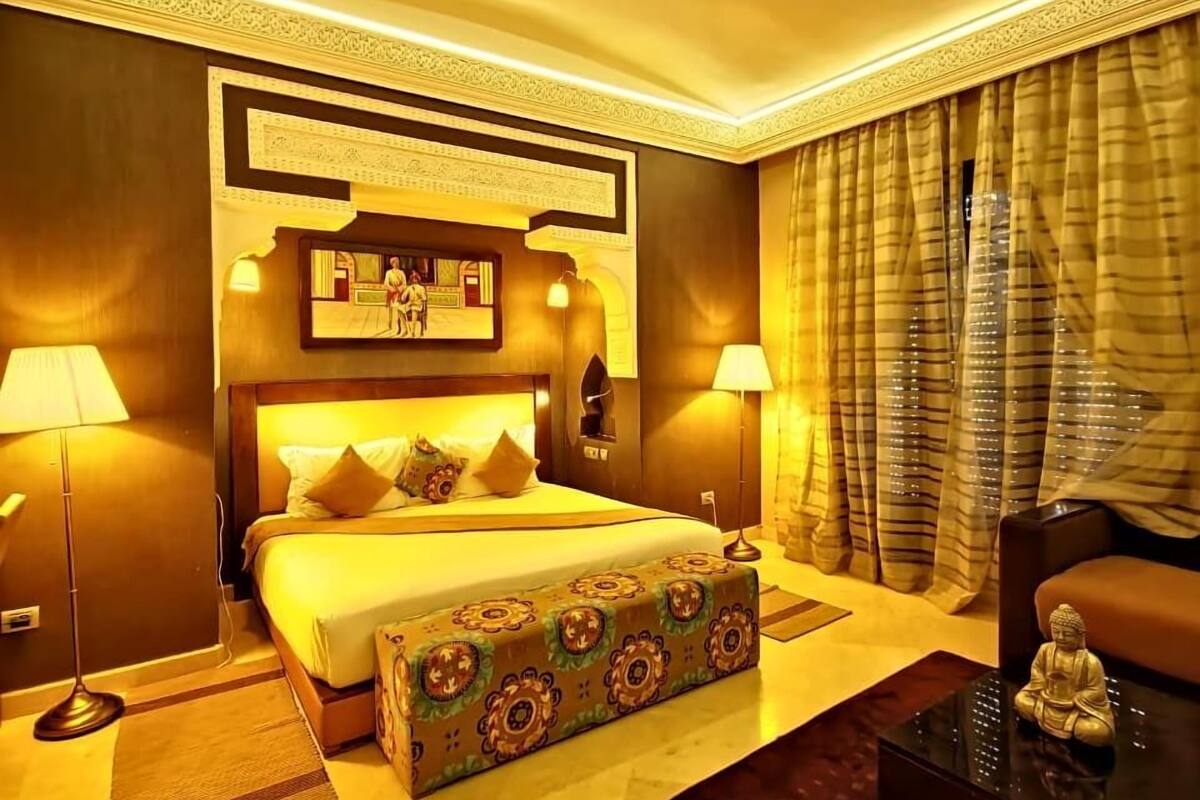 A hotel room with a large bed, two bedside lamps, a small sofa, a decorative Buddha statue, and a large window with curtains.