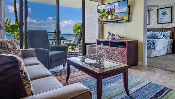 A living room with a sofa, a coffee table, and a TV mounted on the wall. There is a view of the ocean through large windows.