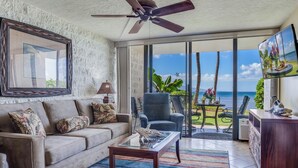 A living room with a sofa, coffee table, and a ceiling fan. There is a large window with a view of the ocean and palm trees.
