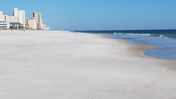 Une vaste plage de sable fin, avec un ciel bleu clair et un océan d’un bleu éclatant. On aperçoit des bâtiments à l’arrière-plan.