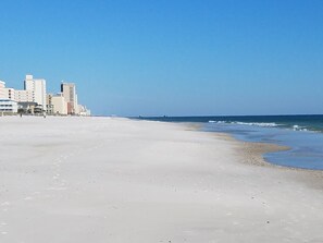 Una amplia playa de arena con cielo azul despejado y mar. Se observan edificios al fondo.