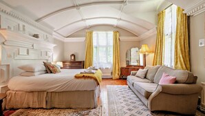 Une chambre avec un grand lit, un canapé, une commode et un miroir. Il y a une fenêtre avec des rideaux jaunes et une lampe sur la commode.