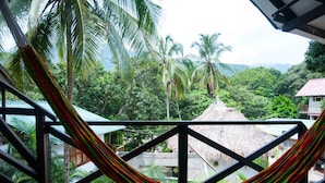 Un hamac sur un balcon donnant sur un paysage luxuriant, avec des palmiers et une construction à toit de chaume.