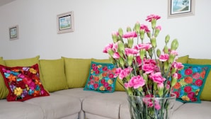 Un salón con un sofá seccional color beige, cojines florales verdes y rojos, y un jarrón con flores rosadas.
