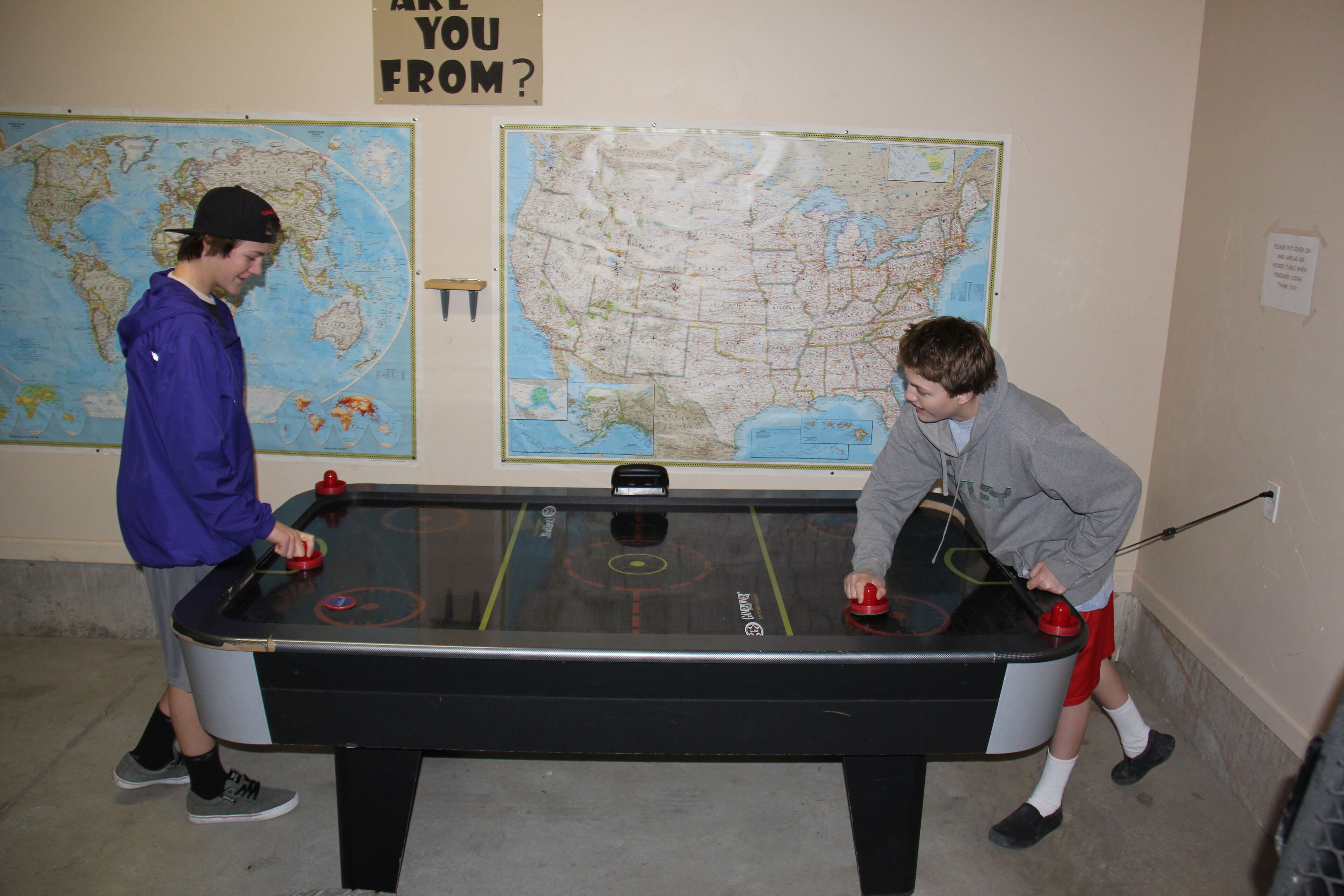Two people playing Foosball in a room with a world map and a U.S. map on the wall.