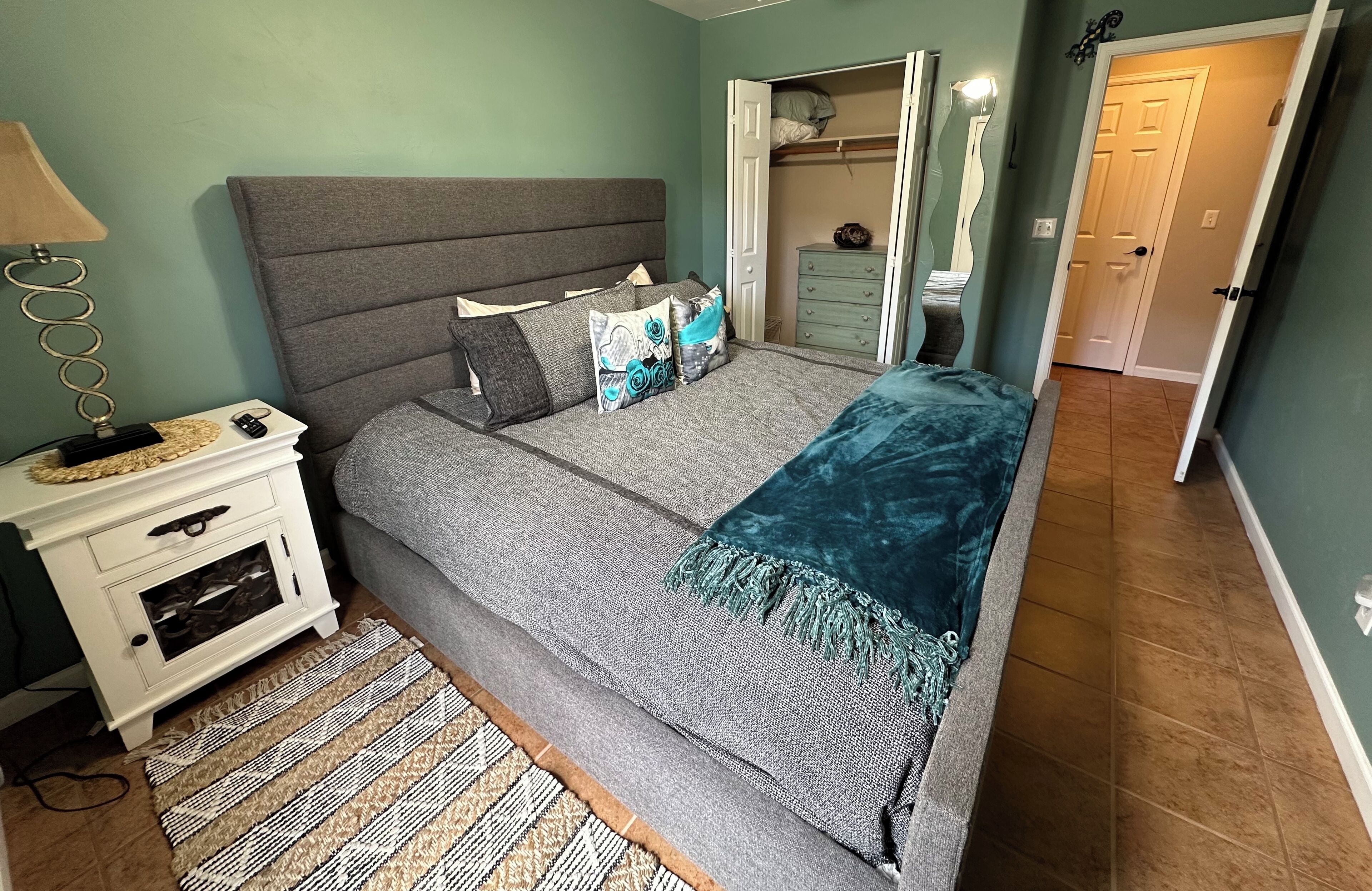 A bedroom with a grey bed, patterned pillows, a bedside table with a lamp, and a woven rug.