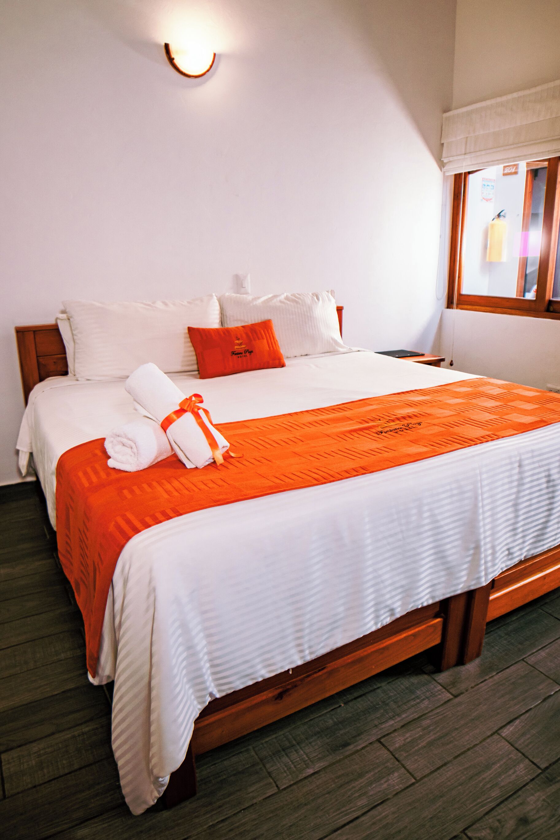 Een net opgemaakt bed met witte en oranje beddengoed, een houten hoofdbord en een aan de muur bevestigde lamp.