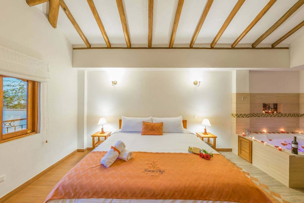 Une chambre d’hôtel avec un grand lit, deux tables de chevet avec des lampes, un plafond en bois, une vue sur un balcon et une salle de bain avec une baignoire et des pétales de rose.