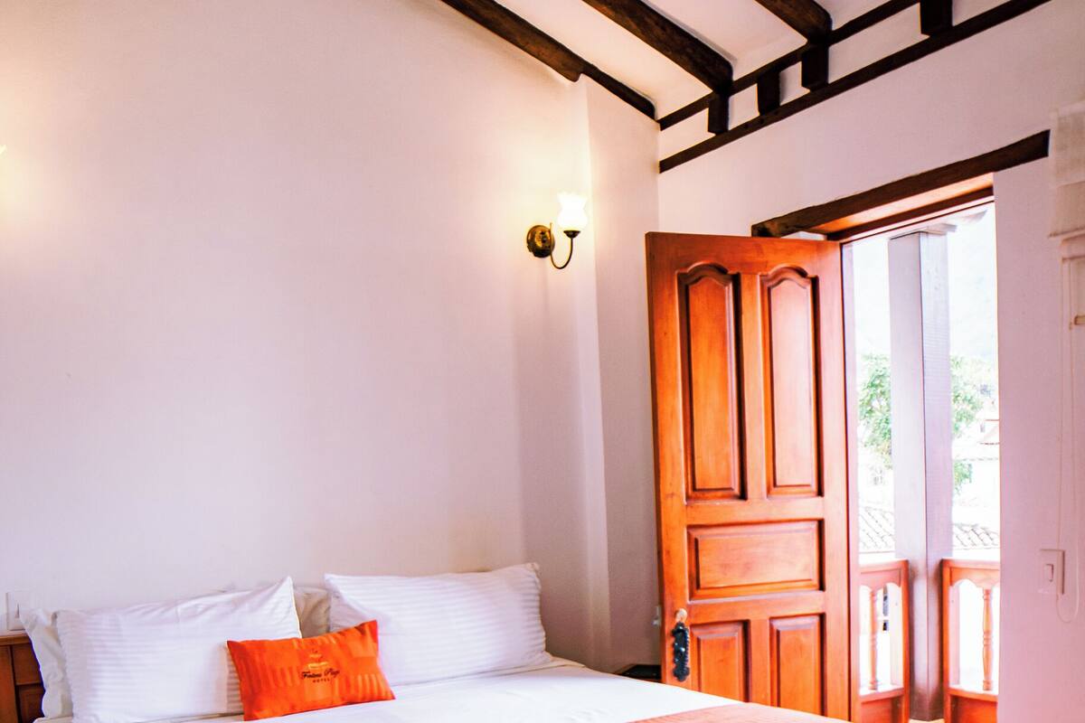 Un lit bien fait, avec une literie blanche et orange, une tête de lit en bois et une porte en bois donnant sur un espace extérieur.
