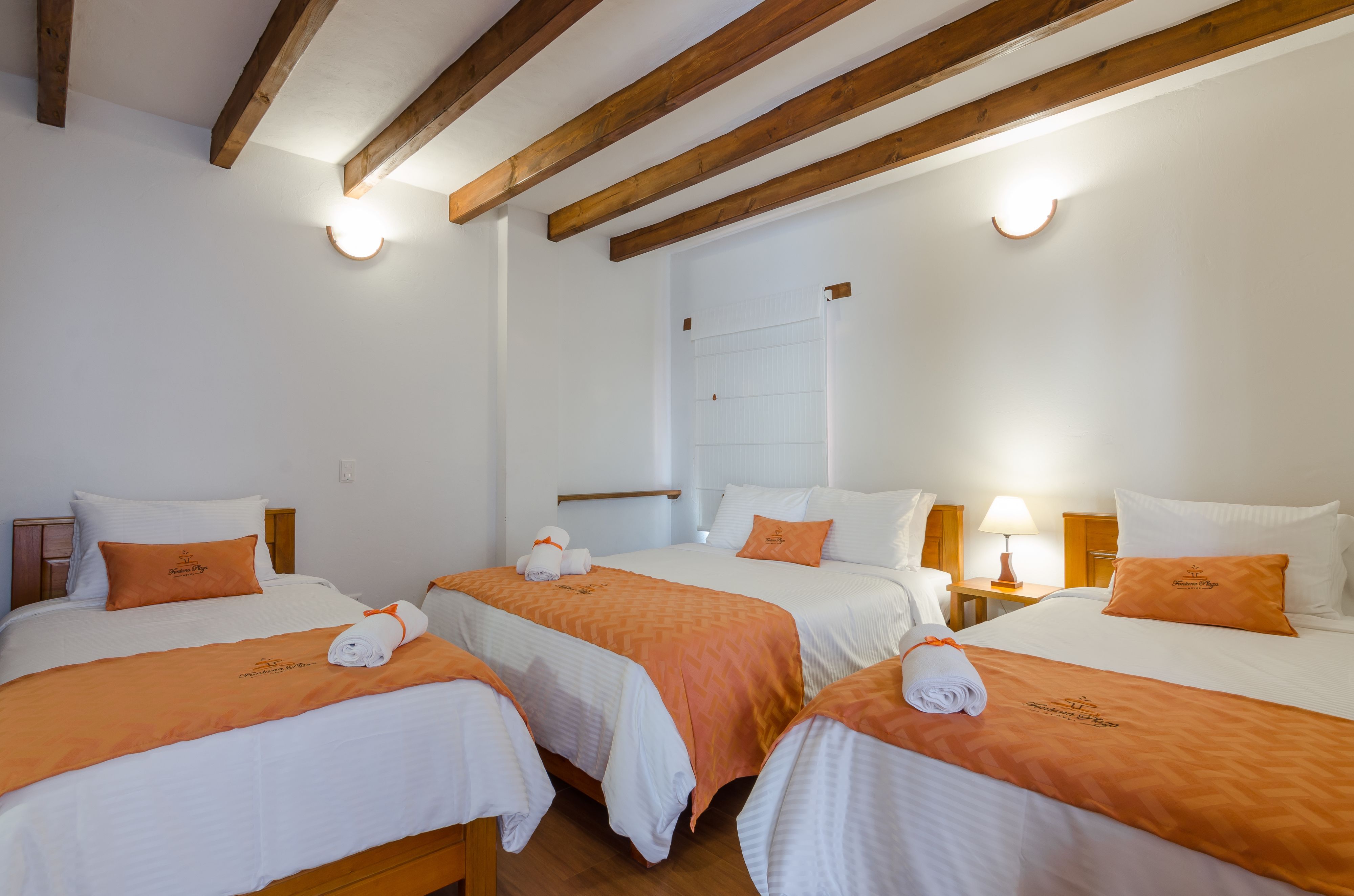 A hotel room with three beds, each with white bedding and orange accents, wooden beams on the ceiling, and a window with blinds.